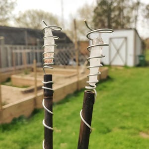 May include: Two handmade crystal wands. Each wand features a clear crystal wrapped in silver wire, attached to a dark brown wooden handle. The wands are set against a blurred outdoor background with a garden and a white shed.