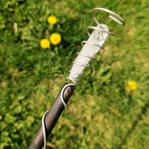 Op de afbeelding: Een handgemaakte toverstaf met een heldere kwartskristal gewikkeld in zilverkleurig draad. De toverstaf heeft een donkerbruine houten handgreep. De achtergrond is wazig groen gras met gele bloemen.