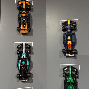 Könnte beinhalten: Vier Miniatur-Rennwagen aus Spielzeug, die an einer grauen Wand ausgestellt sind. Die Autos sind schwarz mit verschiedenen farbigen Akzenten. Das oberste Auto ist orange und schwarz, das zweite Auto ist blau und gelb, das dritte Auto ist blaugrün und schwarz und das unterste Auto ist grün und schwarz.