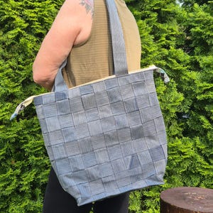 May include: A blue woven tote bag with matching handles and a zip closure. The bag features a basket weave pattern and is carried over the shoulder. The background is a green bush.