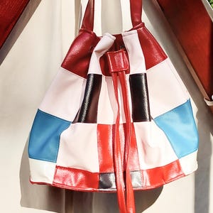 Può includere: Una borsa a secchiello patchwork con chiusura a coulisse. La borsa presenta un vivace mix di colori, tra cui rosso, bianco, nero, blu e rosa. I cinturini e il cordino sono rossi. La borsa è realizzata in materiale lucido.