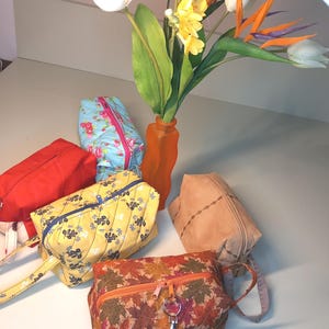 May include: A collection of handmade zippered pouches in various colors and patterns. The pouches include a red pouch, a light blue floral pouch, a yellow floral pouch, a brown leaf-patterned pouch, and a tan suede pouch. A vase of flowers is in the background.