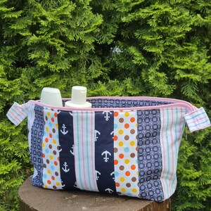 Könnte beinhalten: Eine Kosmetiktasche im Patchwork-Stil mit einem rosa Reißverschluss und gestreiften Griffen. Die Tasche weist verschiedene Stoffmuster auf, darunter marineblaue Anker, Tupfen und geometrische Designs. Zwei weiße Flaschen sind im Inneren der offenen Tasche sichtbar.