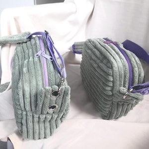 May include: Two corduroy handbags in a soft green colour. Each bag features a purple zip and strap, with silver-toned hardware. The bags have a rectangular shape and a ribbed texture, suggesting a casual style. One bag has a small pocket.