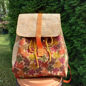 Könnte beinhalten: Rucksack mit Klappenverschluss und orangefarbenem Riemen. Die Klappe und die Seiten bestehen aus hellbraunem Korkmaterial. Die Vorderseite der Tasche zeigt ein farbenfrohes Herbstblattmuster in Rot-, Orange- und Gelbtönen.
