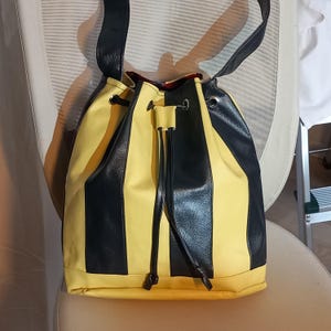 May include: A yellow and black striped bucket bag with a drawstring closure. The bag has a black leather strap and a red patterned interior. The bag is made of a durable material and is designed to be carried over the shoulder.