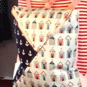 May include: A backpack with a beach theme. The main fabric features a pattern of small beach huts in various colors. The backpack has pink straps and a navy blue section with white anchor designs. The person wearing the backpack is wearing a red and white striped shirt.