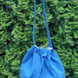 May include: A blue drawstring bag with a long strap, held up against a backdrop of green foliage. The bag is made of a textured fabric and has a drawstring closure. The bag's strap is also blue.