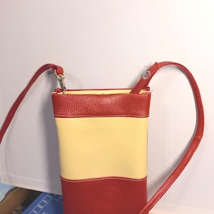 Puede incluir: Un bolso bandolera vertical rectangular con una correa roja y un diseño de bloques de color. Las secciones superior e inferior son rojas, con un centro amarillo. El bolso tiene un cierre plateado y una esterilla de corte en el fondo.