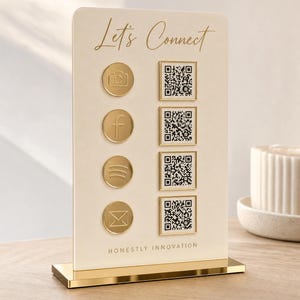 May include: An oatmeal-colored rectangular sign with a gold mirror base featuring the text Let's Connect and HONESTLY INNOVATION. It displays four circular gold social media icons and four corresponding square QR codes in gold frames.