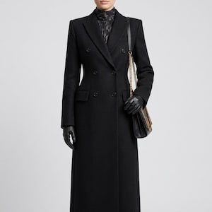 May include: A long, black double-breasted coat with a tailored fit. The coat features a classic notched collar, two rows of buttons, and a sleek, streamlined silhouette. The coat is paired with black gloves and a black shoulder bag with a gold chain.