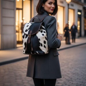 May include: A stylish backpack with a cowhide pattern in black and white, featuring a brown leather flap and straps. The backpack is worn over a gray coat, suggesting a fashion accessory.