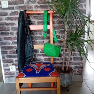 Peut inclure: Chaise en bois avec cadre et pieds orange, assise à motifs bleus. Une veste en cuir noir est suspendue au dossier et un sac à main vert est posé sur le dessus. Une plante en pot se trouve à droite contre un mur de briques.