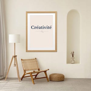 May include: A framed print with the word "Créativité" in navy blue lettering. The print is in a light wood frame and hangs on a cream-colored wall. A woven chair and a small woven ottoman are in front of the wall.
