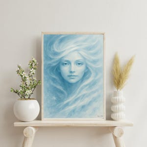 Peut inclure: Une impression d'art encadrée représentant une femme aux cheveux bleus clairs et fluides. L'œuvre d'art a une qualité douce et éthérée, avec une palette de couleurs bleu clair et blanc. L'impression est exposée sur une étagère en bois clair.