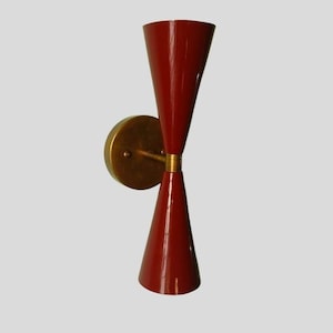 May include: A retro wall sconce featuring a red conical shade and a brass-coloured mounting plate. The light fitting has a double-cone design, with a brass accent in the middle. The sconce is designed to be mounted on a wall.