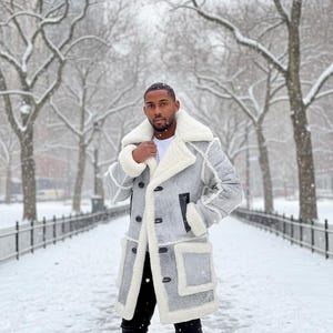 May include: A gray winter coat with a white shearling lining and trim. The coat features a double-breasted closure, black buttons, and two front pockets. The person is wearing black pants and a white shirt. The background is a snowy park.