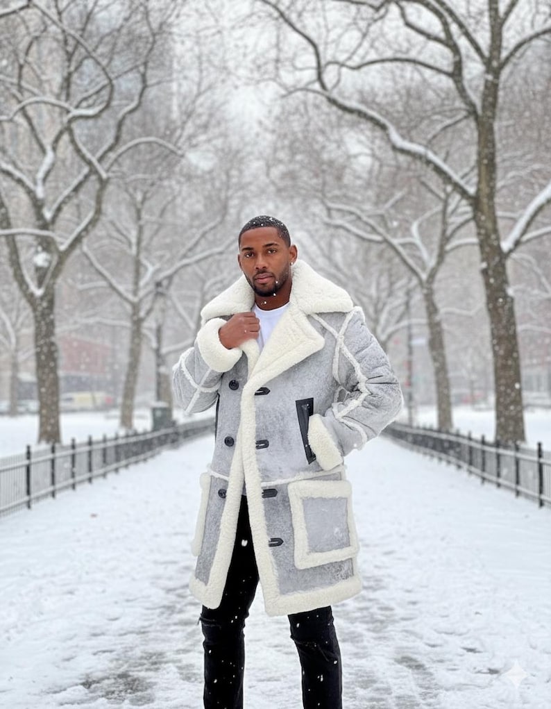 May include: A gray winter coat with a white shearling lining and trim. The coat features a double-breasted closure, black buttons, and two front pockets. The person is wearing black pants and a white shirt. The background is a snowy park.