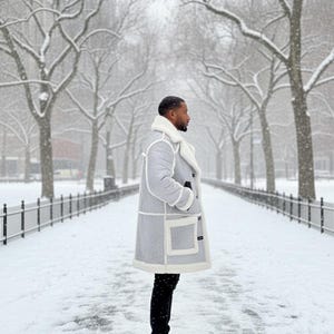 May include: A light gray winter coat with white trim and a white shearling collar. The coat has large pockets and is worn with black pants and boots. The person is standing on a snow-covered path lined with trees.