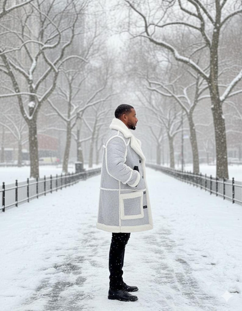 May include: A light gray winter coat with white trim and a white shearling collar. The coat has large pockets and is worn with black pants and boots. The person is standing on a snow-covered path lined with trees.