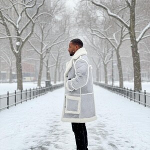May include: A light gray patchwork coat with a white shearling collar and trim. The coat has large square pockets and is worn with black pants and black boots. The background is a snowy park with bare trees.