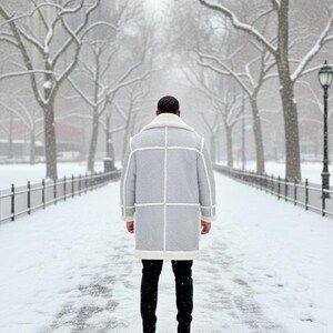 May include: A light gray winter coat with white trim and a white shearling collar. The coat is worn over black pants and black shoes. The person is standing on a snow-covered path in a park with bare trees and a wrought iron fence.