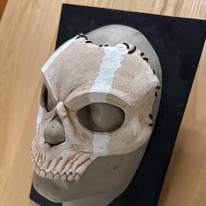 May include: A skull mask in beige and white, mounted on a black base. The mask has stitched details and white stripes. The eye sockets and nasal cavity of the skull are visible. The mask is likely for costume or decorative purposes.
