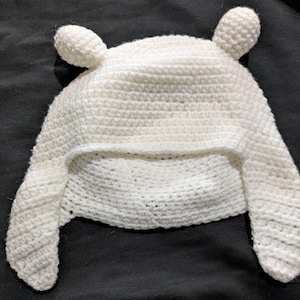 May include: A white crocheted hat with two small, rounded ears on top and ear flaps. The hat is made of a thick, textured yarn and appears to be handmade. The hat is designed to cover the head and ears.