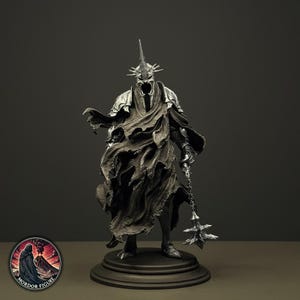Op de afbeelding: Een zilverkleurig beeldje van een lange, gepantserde entiteit met een helm met stekels en een gescheurde, golvende mantel. De figuur houdt een knots met stekels vast en staat op een ronde zwarte voet. Een rond logo bevat de tekst MORDOR FIGURE.