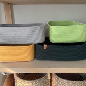 May include: A collection of rectangular storage baskets in various colors, including gray, yellow, green, and teal. The baskets are made of a woven material and are displayed on a wooden shelf. Each basket has a small brown tag.