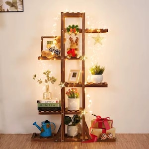 May include: A wooden shelf unit with five shelves, decorated with fairy lights, a teddy bear, a reindeer figurine, a star ornament, and potted plants. The shelf unit is against a white wall.
