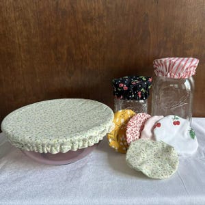 May include: A collection of fabric bowl covers with elastic edges, featuring various patterns including green floral, yellow floral, red cherry, and red stripes. One cover is placed over a glass bowl, while others are stacked or placed on a glass jar.