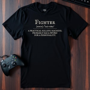 Puede incluir: Camiseta negra con la palabra "Fighter" y su definición impresa en blanco. Un mando de juegos y un teclado negros son visibles sobre una superficie de madera oscura.