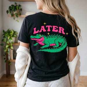 Puede incluir: Camiseta negra con un gráfico de un caimán verde y rosa y la palabra "LATER" en rosa. El diseño incluye acentos de estrellas amarillas. Una prenda de vestir informal.