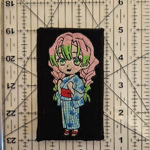 May include: Embroidered patch featuring a cartoon character with pink hair and green accents, wearing a blue kimono. The patch is on a black background. The patch is approximately 2 inches wide and 3 inches tall.