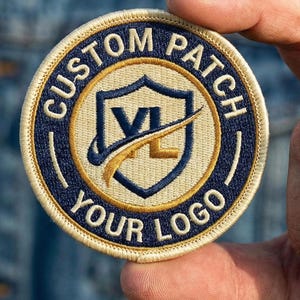 May include: Circular embroidered patch with the text "CUSTOM PATCH YOUR LOGO". The patch features a shield design with the letters "VL" and a swoosh. The patch has a navy blue and gold color scheme.
