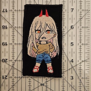 May include: Embroidered patch featuring a cartoon character with blonde hair, red horns, and orange eyes. The character wears a tan shirt, blue shorts, and red shoes. The patch is rectangular with a black background.