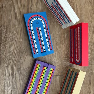 May include: Five different wooden cribbage boards with various colour schemes. The boards are all in the shape of a horseshoe and have a small hole in the centre. The boards are all made of wood and have a smooth finish. The boards are all different colours, including blue, red, purple, yellow, and brown.