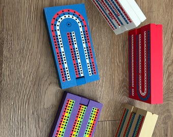 Peut inclure: Cinq plateaux de cribbage en bois avec différentes combinaisons de couleurs. Les plateaux sont tous en forme de fer à cheval et ont un petit trou au centre. Les plateaux sont tous en bois et ont une finition lisse. Les plateaux sont tous de couleurs différentes, y compris le bleu, le rouge, le violet, le jaune et le brun.