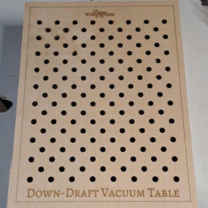 May include: A rectangular wooden down-draft vacuum table with many evenly spaced holes. The table has the text "Down-Draft Vacuum Table" and the "Barnes Woodworks" logo etched into the wood.