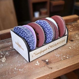 Może przedstawiać: Drewniany organizer na papier ścierny z wycinanym laserowo wzorem, mieszczący kilka okrągłych krążków papieru ściernego w różnych kolorach. Organizer ma wygrawerowane słowa "Sandpaper Organizer" i logo z przodu.