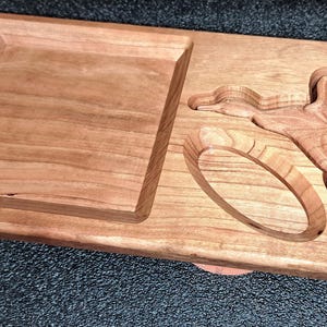 May include: A rectangular wooden serving tray with a carved design. The tray features a square recessed area and a carved flying bird with a ring. The wood has a warm, reddish-brown hue.