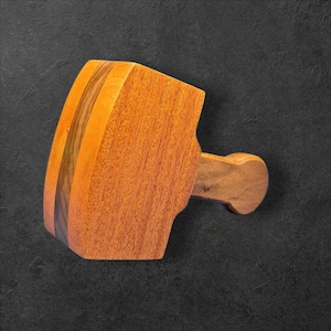 May include: A wooden mallet with a rounded head and a handle. The mallet is made of various shades of wood, including light and dark brown. The handle is attached to the head with a curved design.