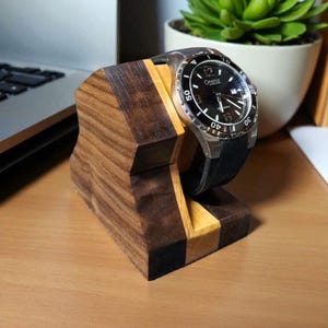 May include: A wooden watch stand with a dark brown and light wood grain design. A black-faced watch with a silver bezel and black band rests on the stand. The watch has white numerals and hands.