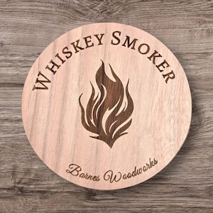 以下が含まれることがあります： 上部に「Whiskey Smoker」の文字が刻まれた丸い木製コースター。中央には炎のデザインがあります。「Barnes Woodworks」の文字が下部に刻まれています。コースターは明るい茶色で、濃い茶色の炎のデザインです。