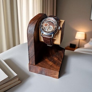 May include: A brown leather-banded wristwatch with a silver case rests on a wooden watch stand. The watch has a brown face with visible inner workings. The stand is made of dark brown wood and has a unique, angled design.