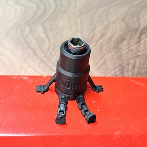 May include: A black 10mm socket wrench with arms and legs, giving it a cartoon-like appearance. The wrench is sitting on a red surface, with a wooden background.