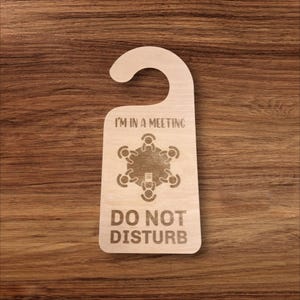 以下が含まれることがあります： 「I'M IN A MEETING」の文字と、テーブルを囲む人々のグラフィックが描かれた木製のドアハンガー。「DO NOT DISTURB」の文字も含まれています。ハンガーは明るい茶色で、ドアノブに掛けるためのカーブした上部があります。