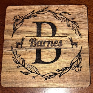 Puede incluir: Posavasos cuadrado de madera con un diseño decorativo. El posavasos presenta una gran letra "D" con el nombre "Barnes" debajo. Una corona floral y siluetas de ciervos rodean la letra. La madera tiene una veta natural.
