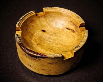 May include: A handcrafted wooden ashtray with a natural, light-yellow finish. The round ashtray features three notched rests for cigarettes and a dark, reddish-brown accent on the rim. The wood grain is visible, adding to its rustic charm.